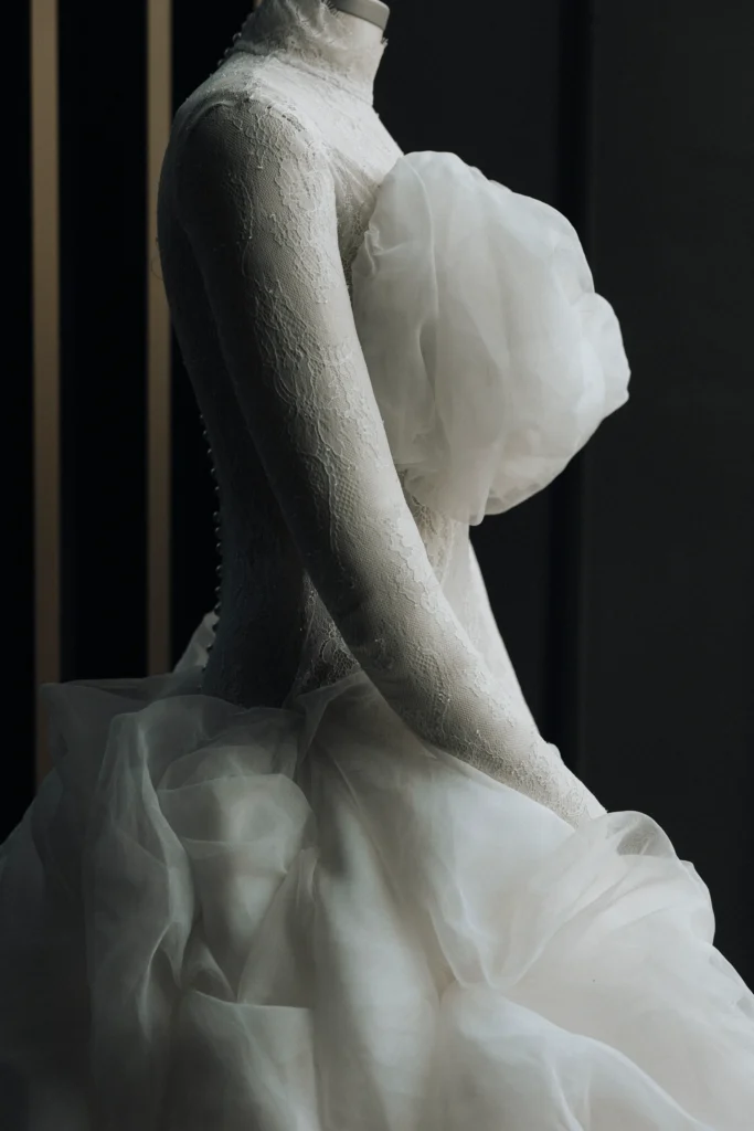 A white lace wedding dress with sheer fabric details is displayed on a dress form against a dark background.